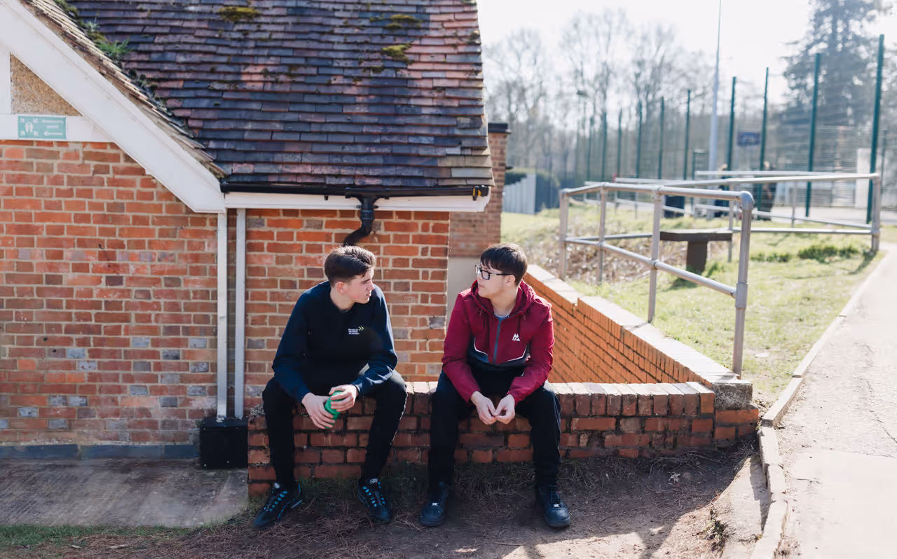 Two young men sitting on a brick ledge outside, engaged in conversation near a brick building.