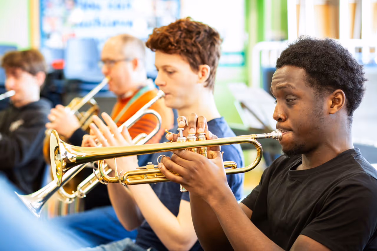Four male trumpet players sitting in a row, focused on playing their instruments indoors.