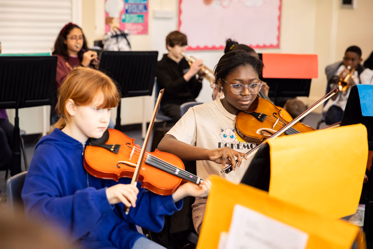 Two children playing violins in a classroom orchestra with other students playing wind instruments in the background.
