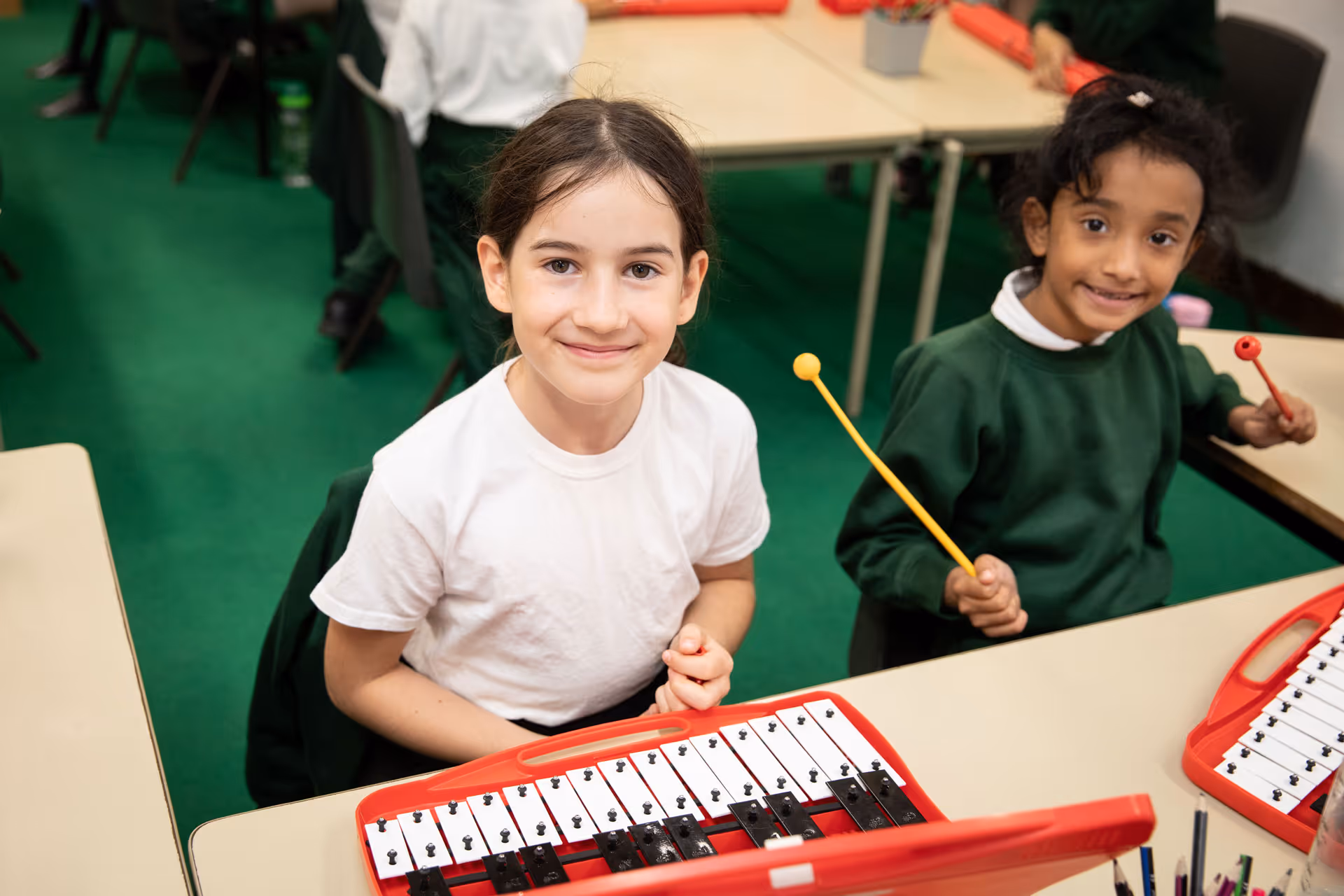 Two young girls sitting at desks playing red xylophones with mallets, smiling at the camera.