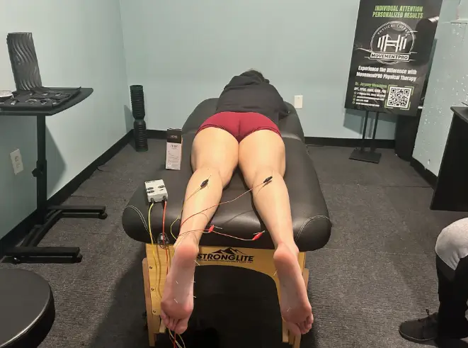 Person lying face down on a therapy table with electrodes attached to their calves for physical therapy treatment.