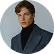 Portrait of a young man with light brown hair wearing a black blazer and a dark shirt against a plain gray background.