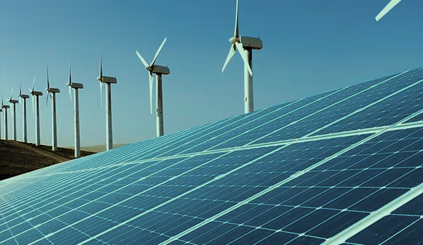 Solar panels in the foreground with a row of wind turbines in the background under a clear blue sky.