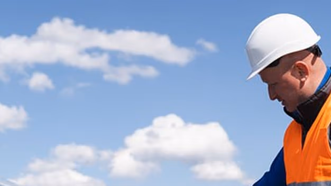 Construction worker wearing a white hard hat and orange safety vest looking downward against a blue sky with scattered clouds.