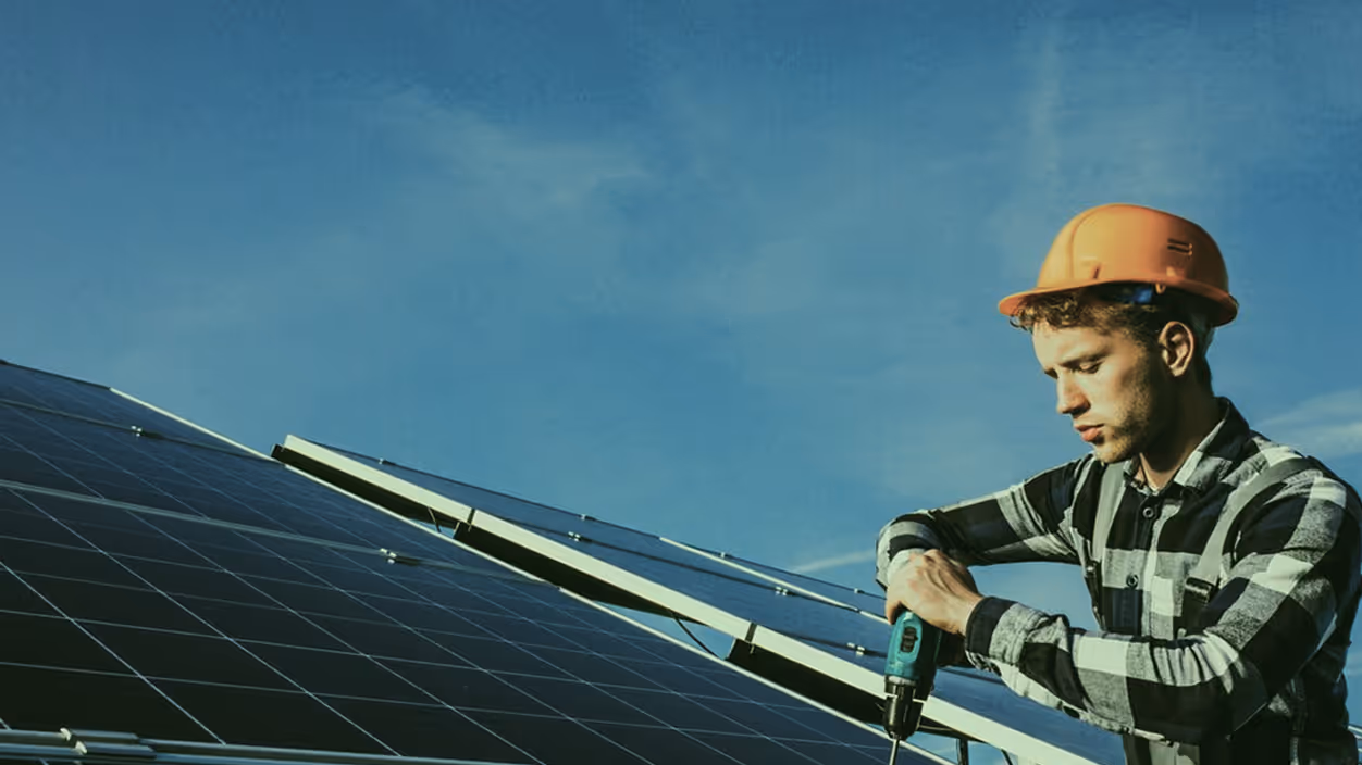 Man wearing an orange safety helmet and plaid shirt installing solar panels with a power drill under a clear blue sky.