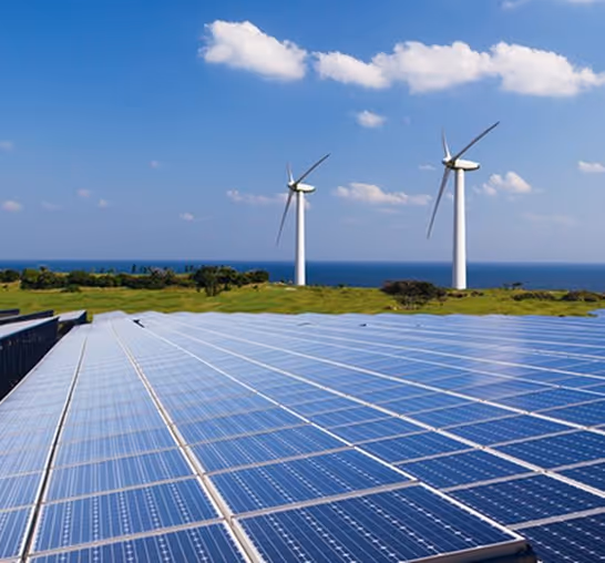 Solar panels in the foreground with two wind turbines and blue sky above near the coast.