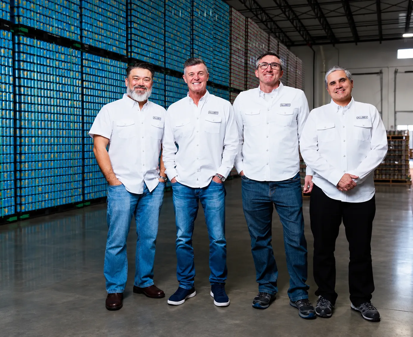 Four men in white button-down shirts and jeans stand in a warehouse with stacks of cans in the background.