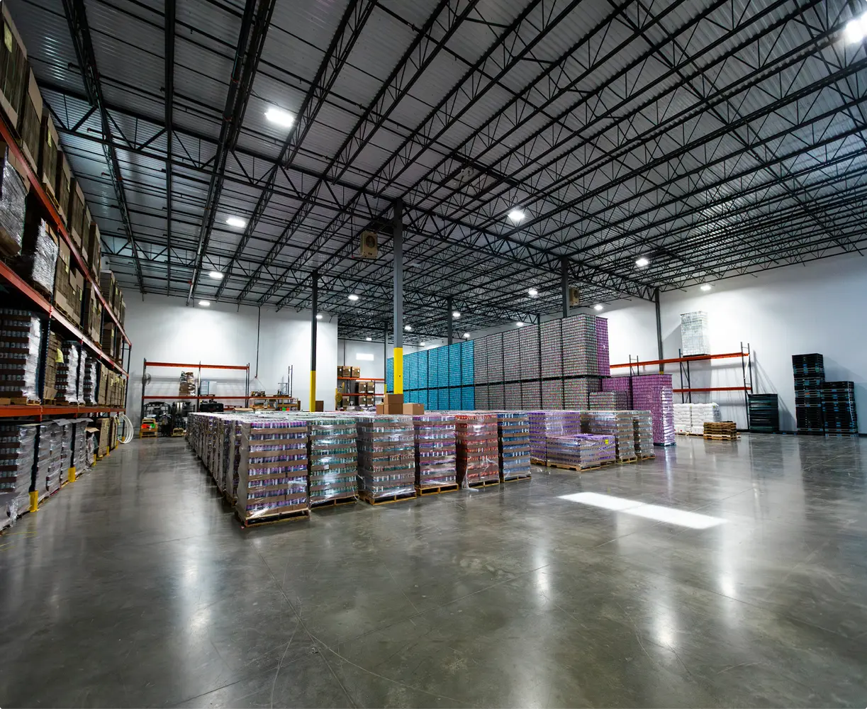 Pallets stacked with colorful beverage cans are arranged in rows inside a large, well-lit warehouse with a high ceiling.