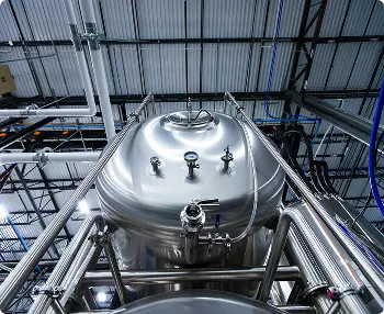 Large stainless steel industrial tank with gauges and pipes inside a modern facility, viewed from a low angle against a metal ceiling.