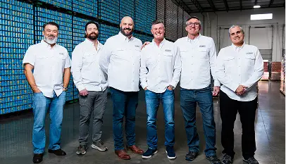 Six men in white button-down shirts and jeans stand in a warehouse with stacks of cans in the background.