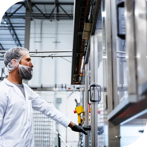 A worker wearing a hairnet and mask operates a control panel on industrial machinery inside a factory.