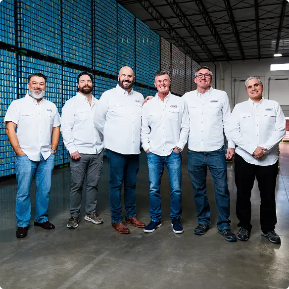 Six men in white button-down shirts and jeans stand in a warehouse with stacks of cans in the background.