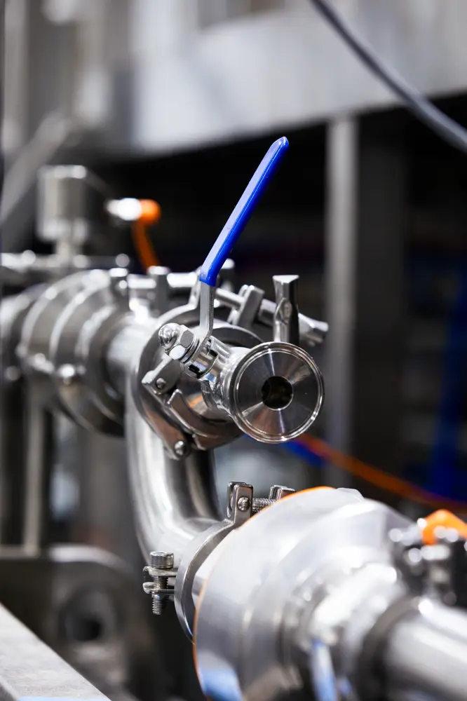 Close-up of a stainless steel industrial valve with a blue handle, attached to a network of metal pipes in a factory setting.