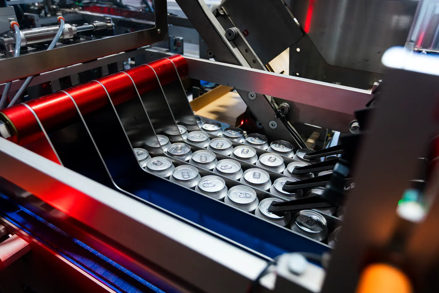 Aluminum cans move along a conveyor belt inside a beverage production facility, surrounded by machinery and metal components.