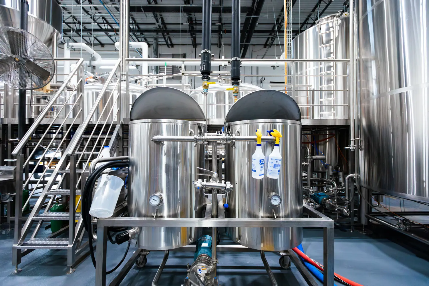 Two large stainless steel tanks with gauges and cleaning bottles are shown in an industrial brewery or food processing facility, with pipes, stairs, and more tanks in the background.