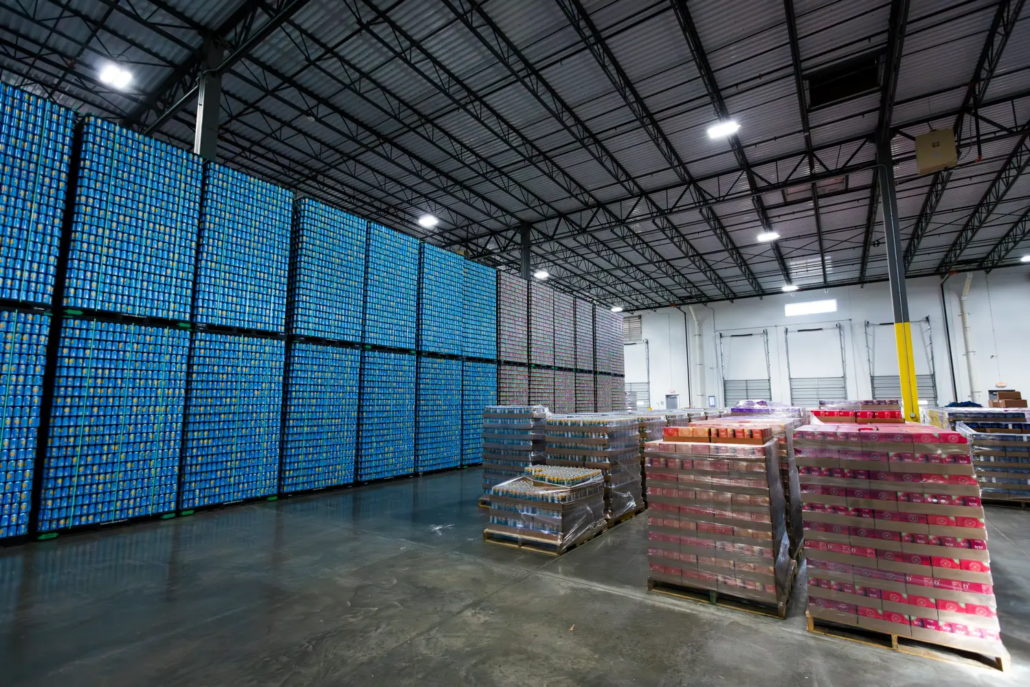 Stacks of aluminum cans and pallets of packaged beverages are arranged in a large warehouse with high ceilings and concrete floors.
