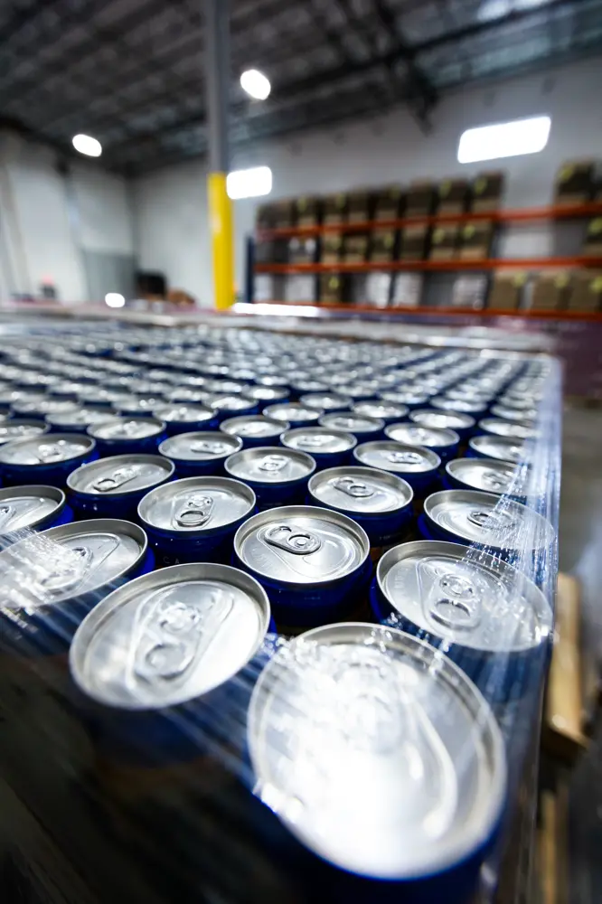 Rows of canned beverages covered in plastic wrap are stacked on a pallet inside a large warehouse with shelves in the background.