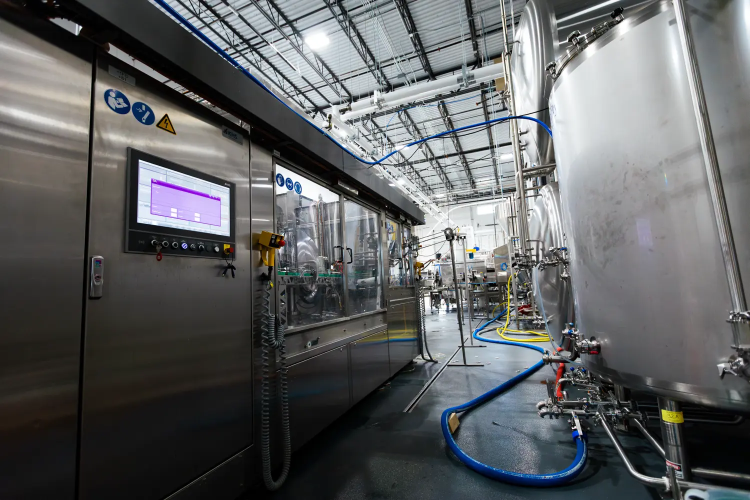 Stainless steel industrial tanks and machinery inside a modern factory with pipes, cables, and a control panel visible.