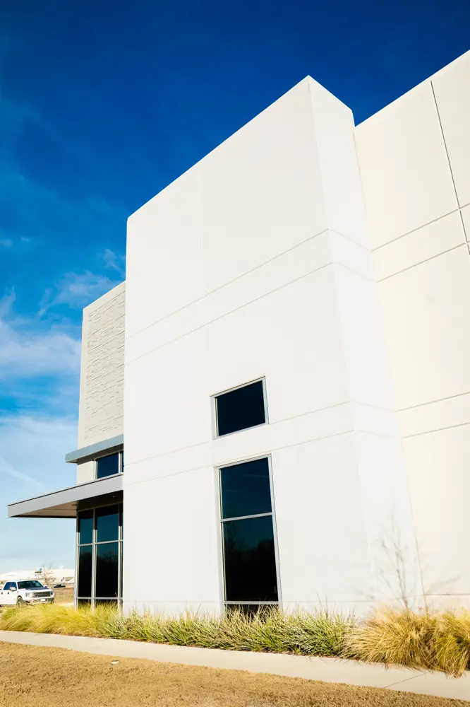 Modern white commercial building with large rectangular windows, clear blue sky, and dry grass landscaping in the foreground.