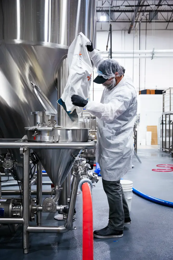 A worker in protective clothing pours ingredients into a large stainless steel tank at an industrial food or beverage processing facility.