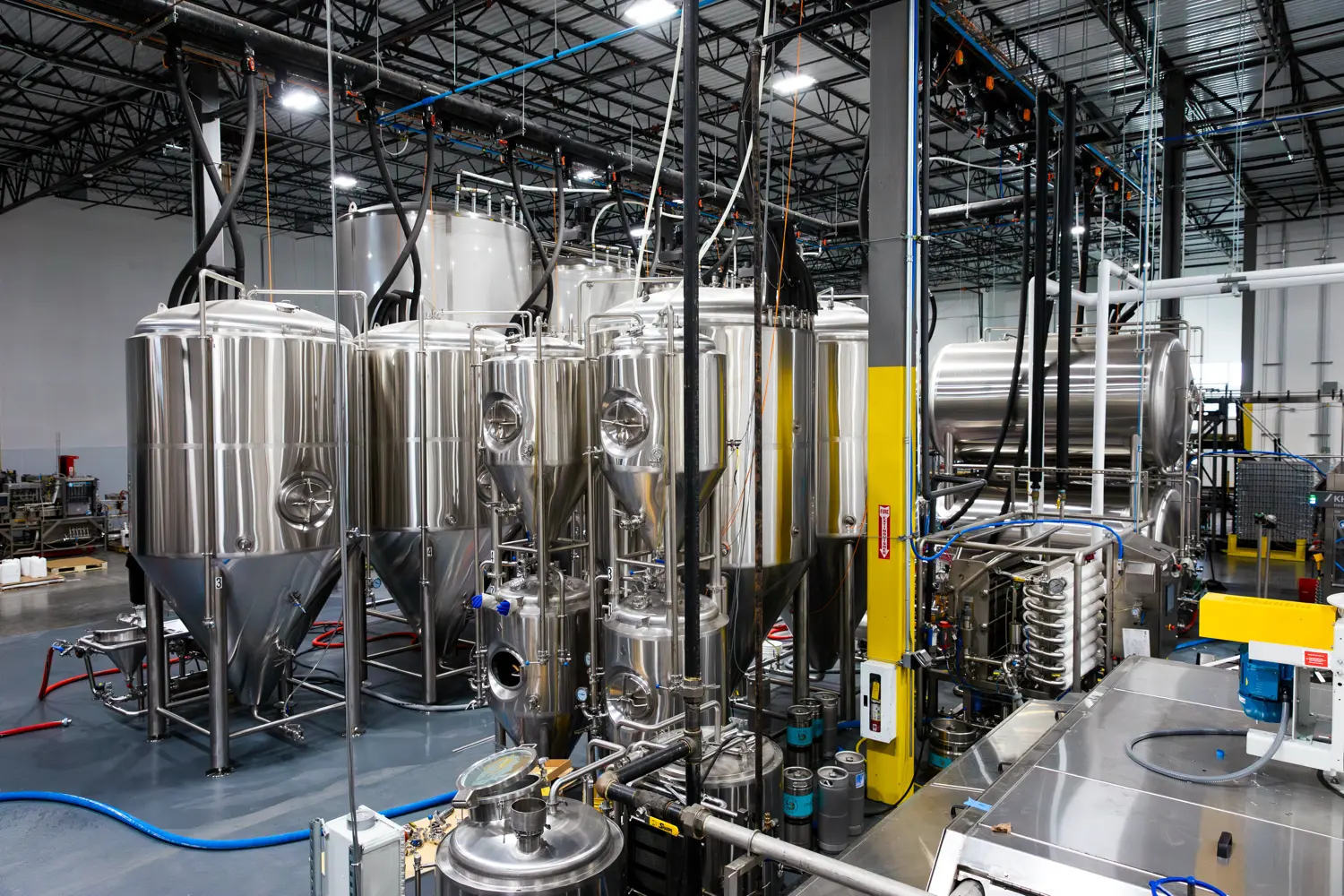 Rows of large stainless steel brewing tanks and pipes inside a modern brewery facility with various equipment and machinery.