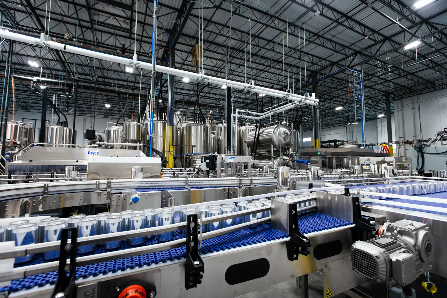 A modern beverage factory interior showing automated canning equipment, conveyor belts with empty cans, and large stainless steel tanks in a spacious, well-lit industrial setting.