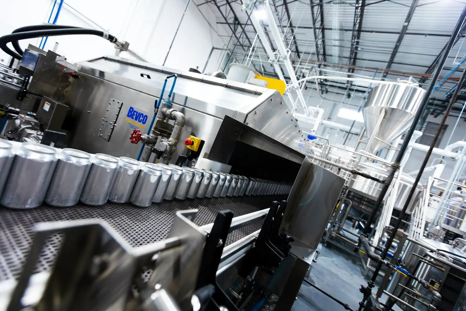 A factory production line with metal cans moving along a conveyor belt, surrounded by industrial machinery and stainless steel equipment.