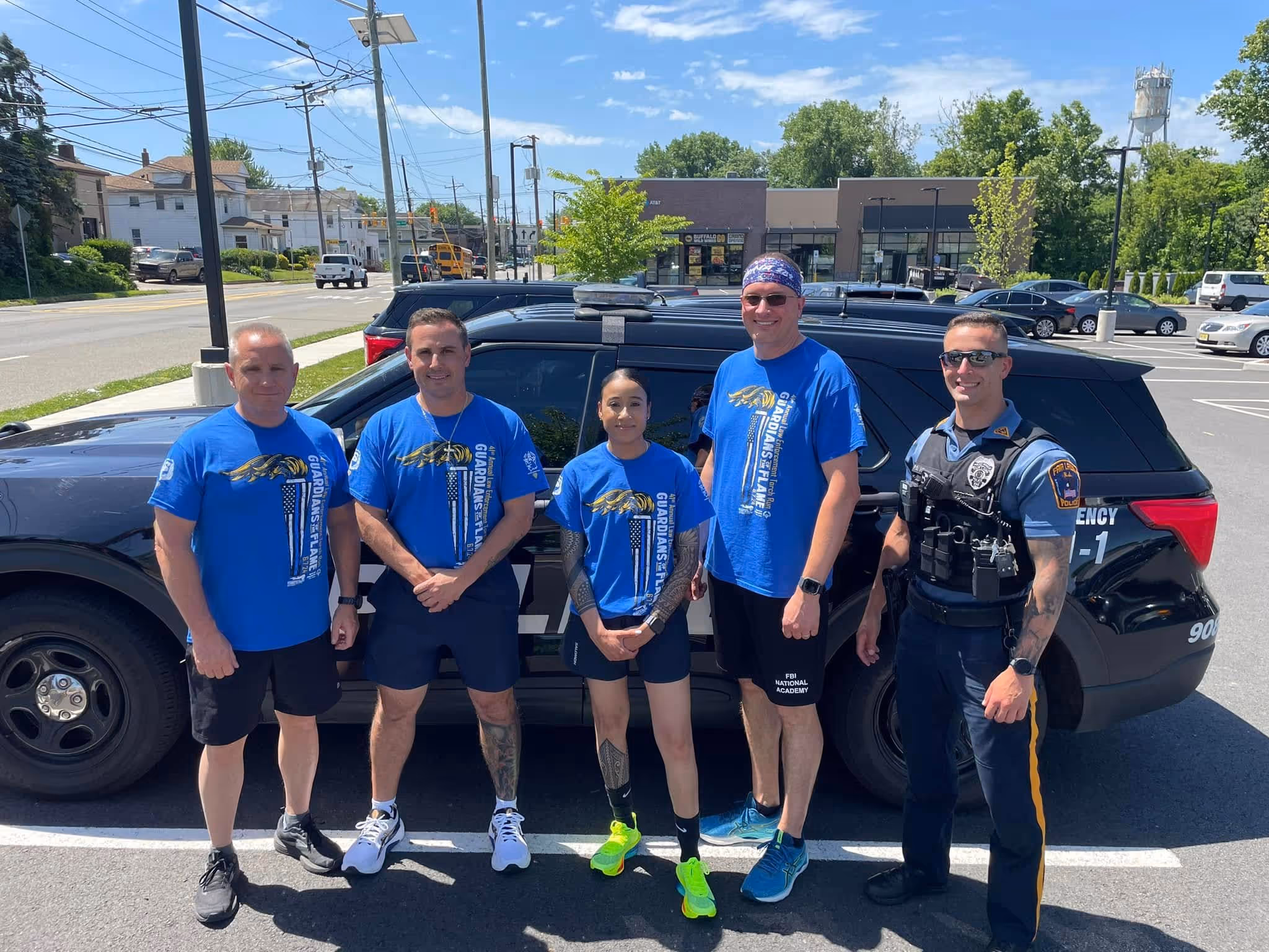 Five people standing in front of a black police SUV, four wearing blue Guardian’s Flame shirts and one in police uniform on a sunny day.
