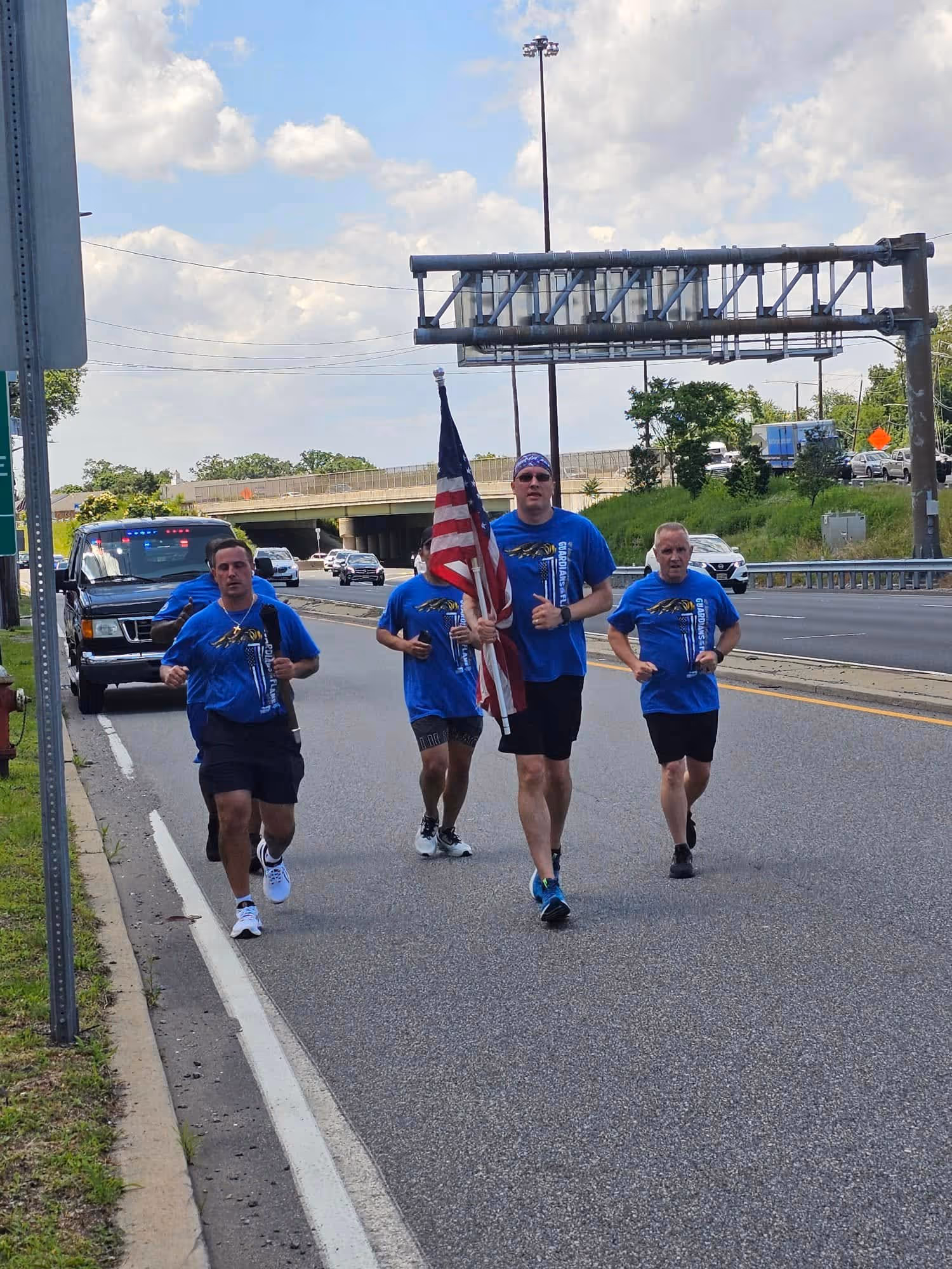 Group of men in blue shirts running on a highway, one holding an American flag, with a police car behind them.