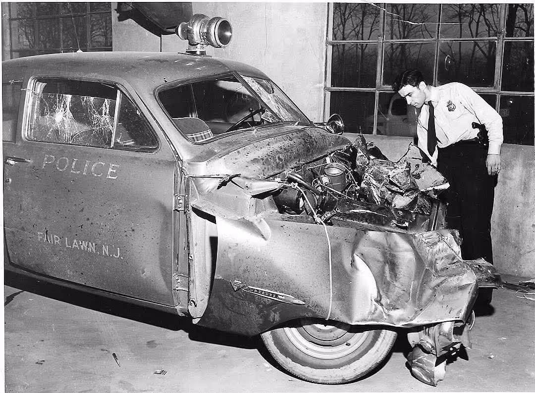 Police officer inspects heavily damaged front of a Fair Lawn, N.J. police car with shattered passenger window.