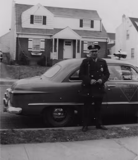 Black-and-white photo of a police officer standing in front of a vintage police car parked on a residential street with houses in the background.