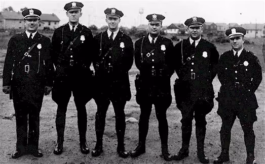 Black and white photo of six police officers in uniform standing outdoors on dirt ground.