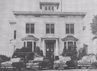Black and white photo of an old Fire Department building with four vintage fire trucks parked in front.