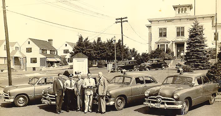 Vintage black and white photo showing six men standing between three classic cars parked on a street with houses and a large building in the background.