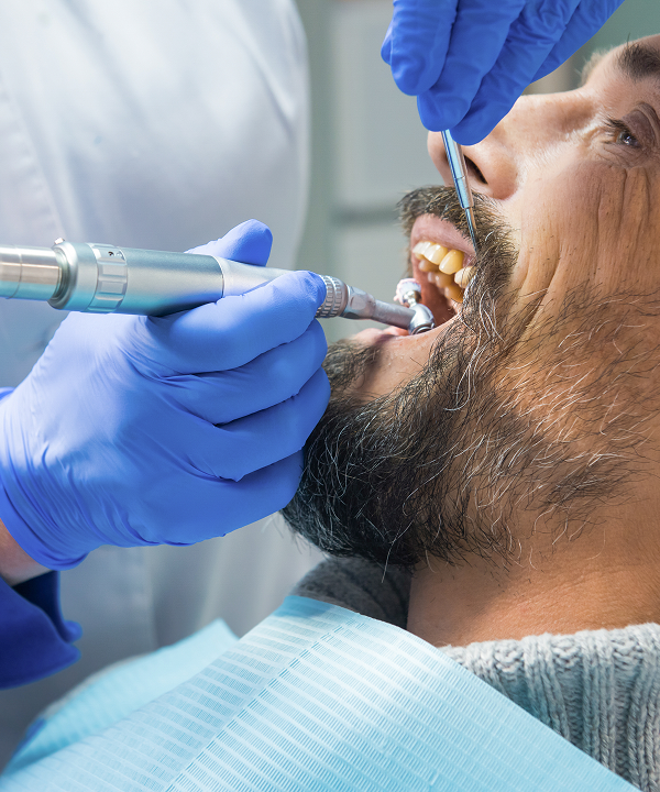 Dental professional wearing blue gloves using dental tools to clean a male patient's teeth.