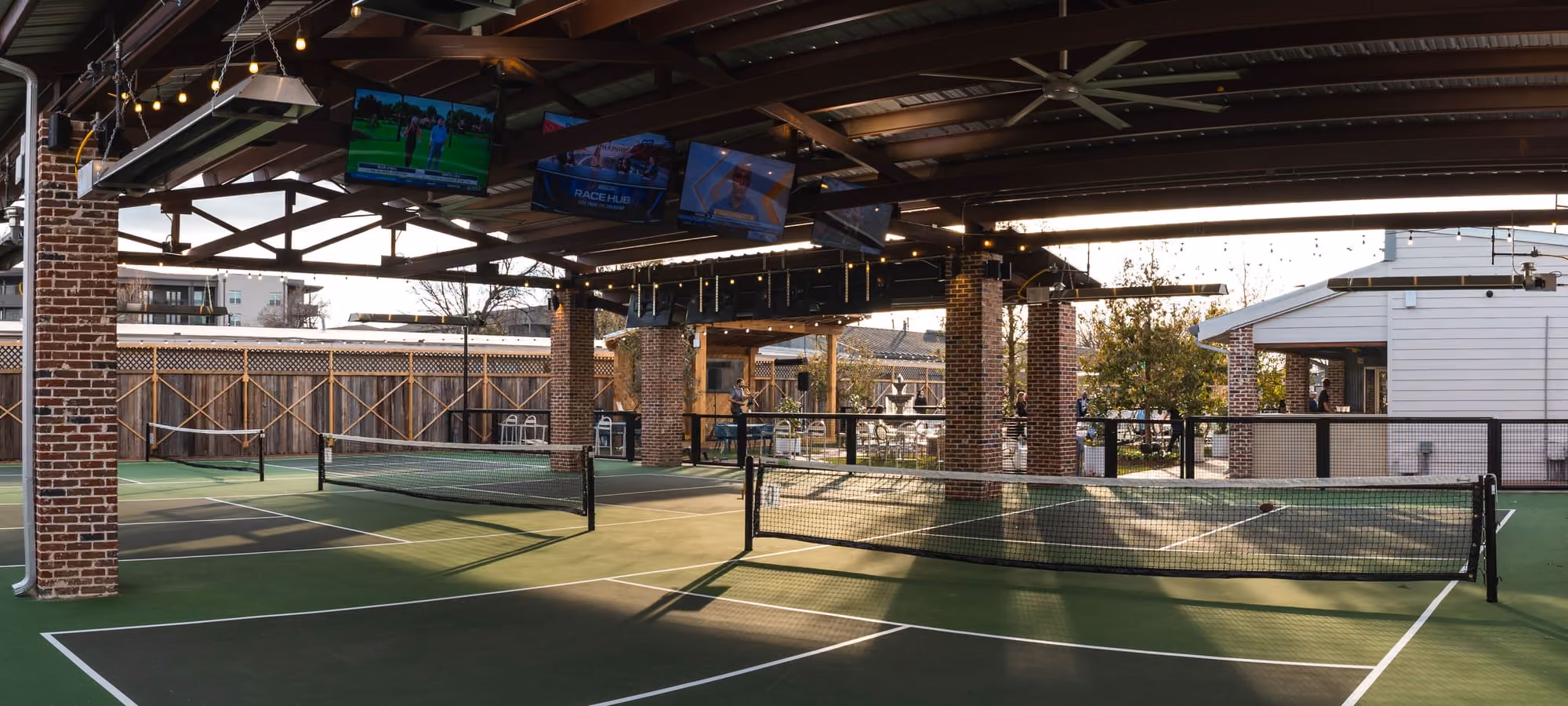 The Courts at Meraki offer a covered pickleball setup with string lights, big-screen TVs, and lawn views beyond.