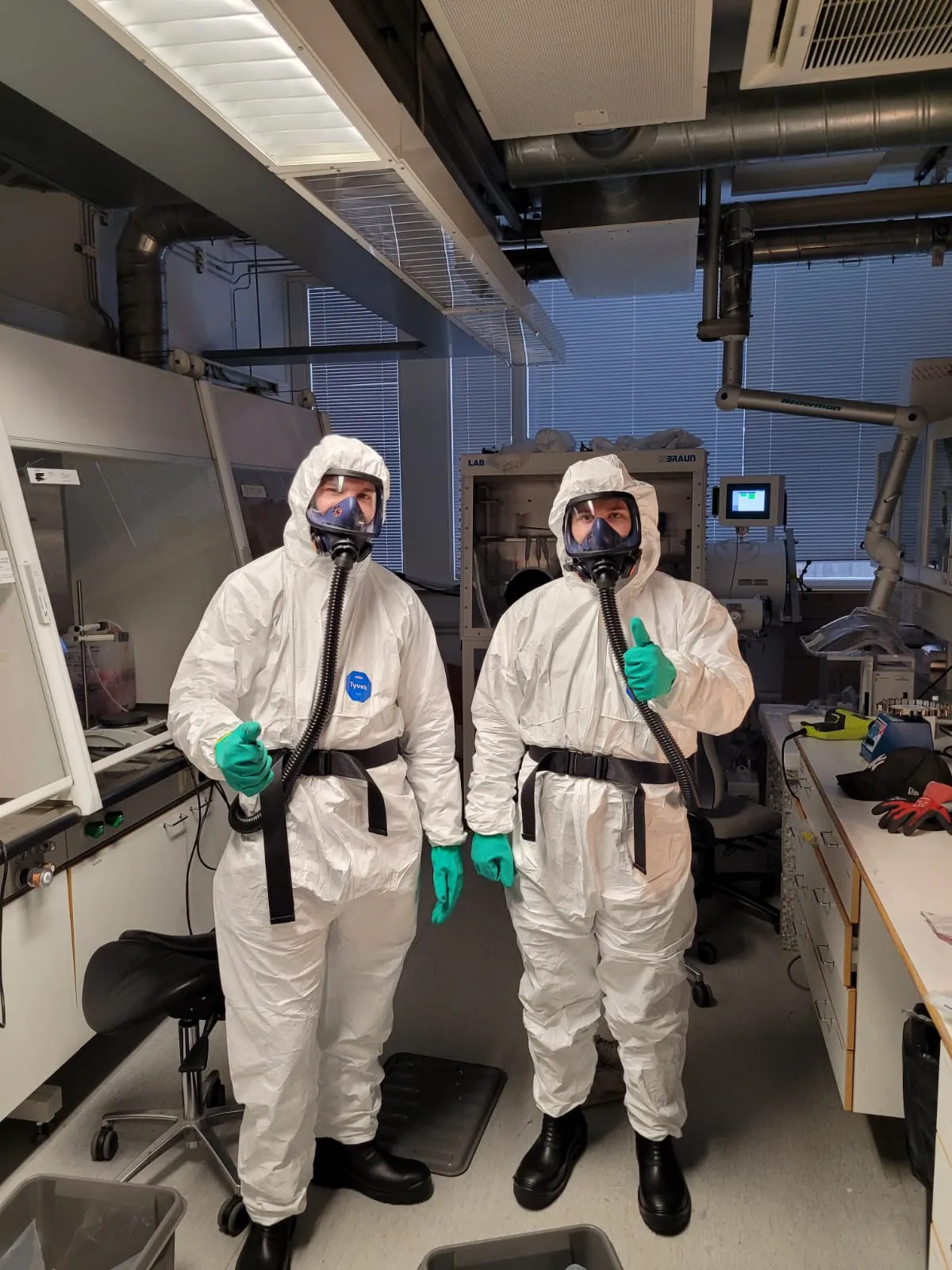 Kaksi henkilöä valkoisissa suojapuvuissa ja hengityssuojaimissa seisoo laboratoriossa, toinen peukalo ylhäällä, kirkkaassa valaistuksessa.