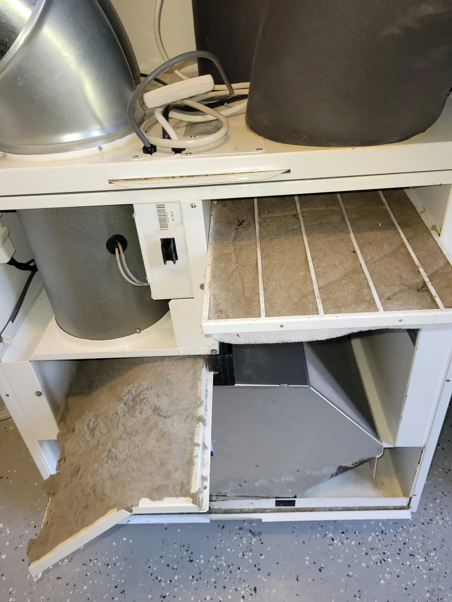 Likaiseen valkoiseen kaappiin on asennettu likainen suodatin, ja kaapin alaosassa on suuri määrä pölyä. Kaapin yläosassa on kaksi metallista sylinteriä.