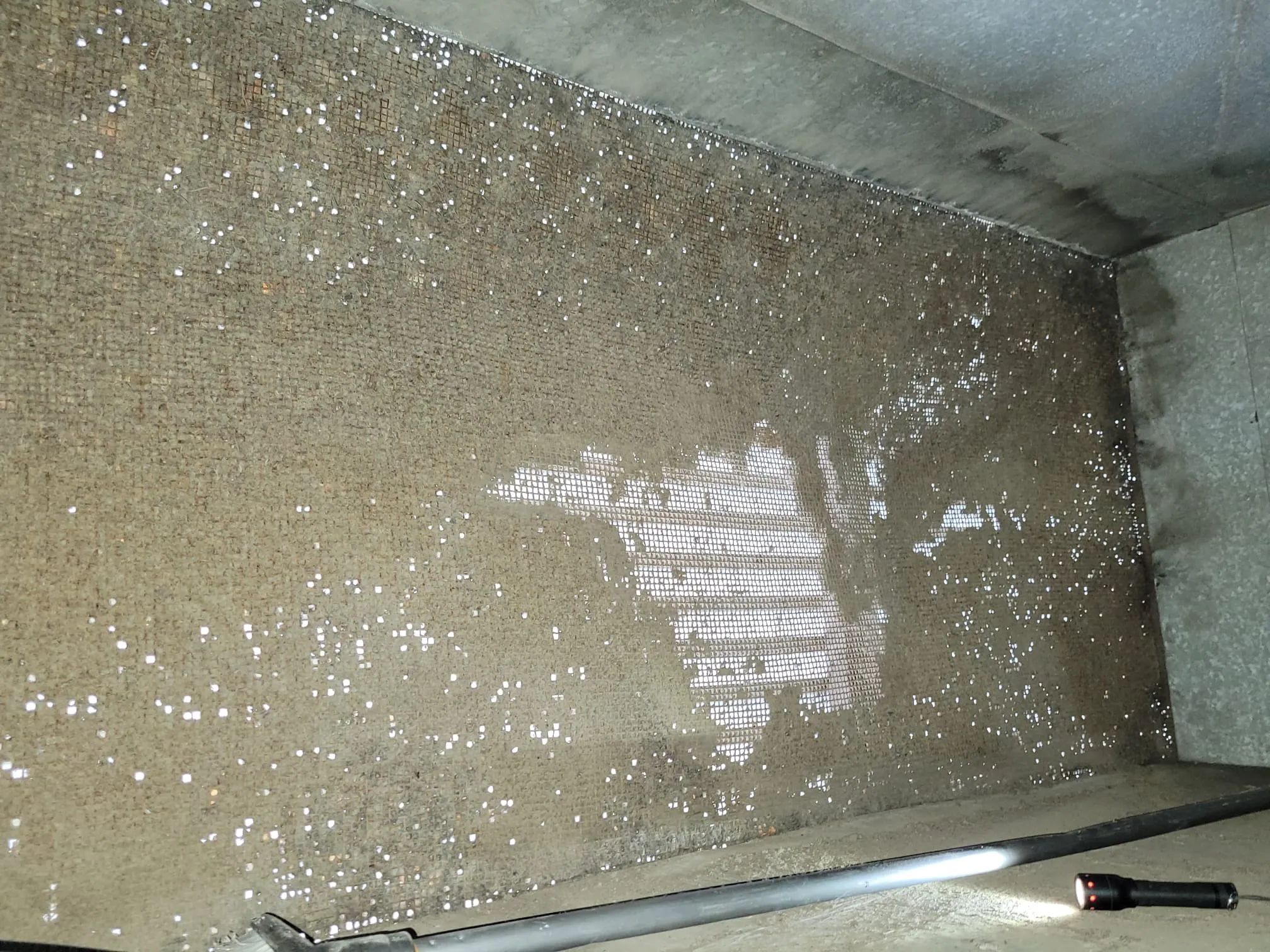 Karkean seinän ja katon sisätila, jossa on metalliverkko ja valkoisia pisteitä, sekä lattialla putki ja taskulamppu.