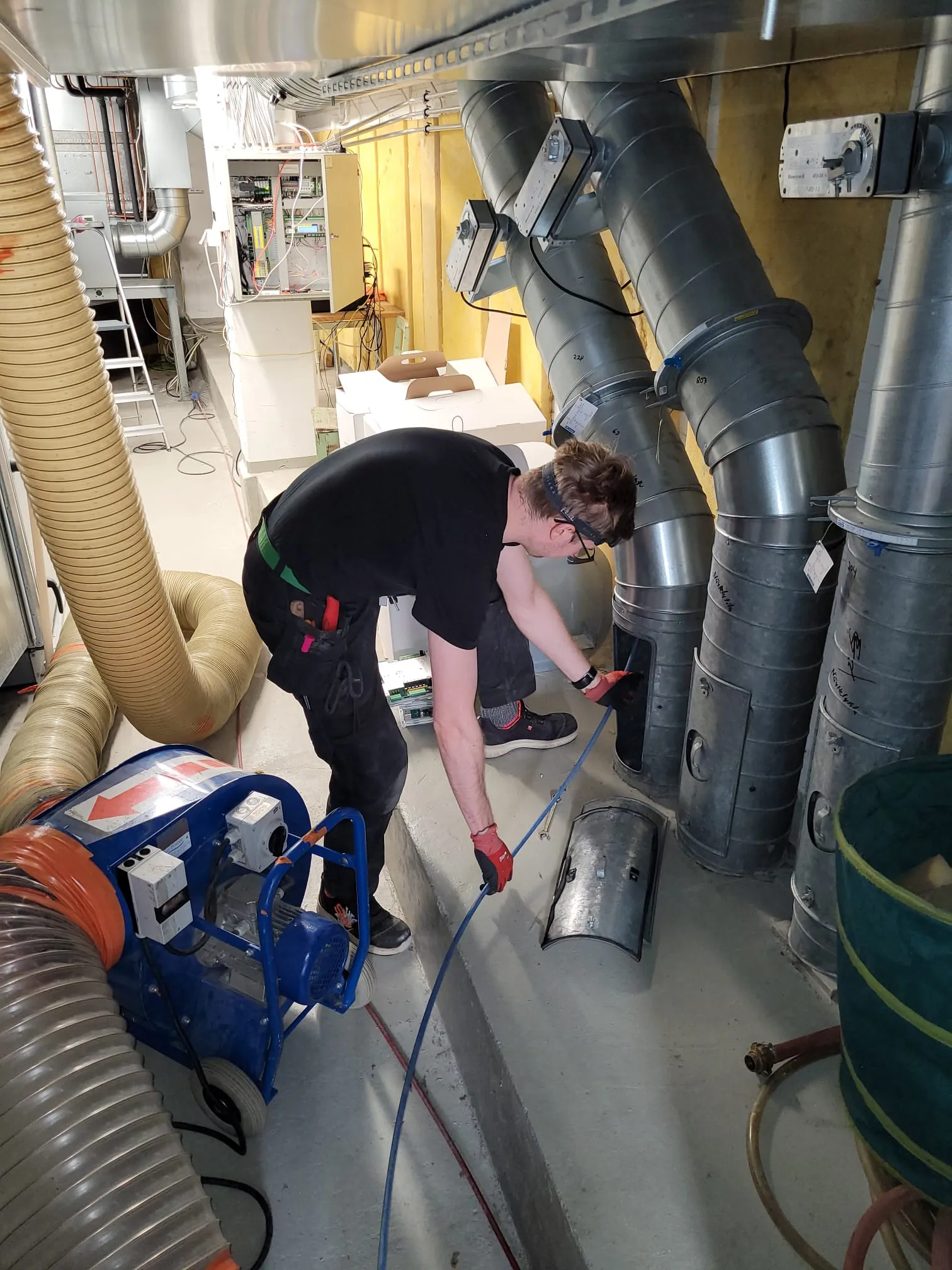 Mies työskentelee ilmastointiputkien parissa, käyttäen suojalaseja ja punaisia työkäsineitä, sininen puhallin ja keltainen putki näkyvät vasemmalla, valaistus on kirkas.