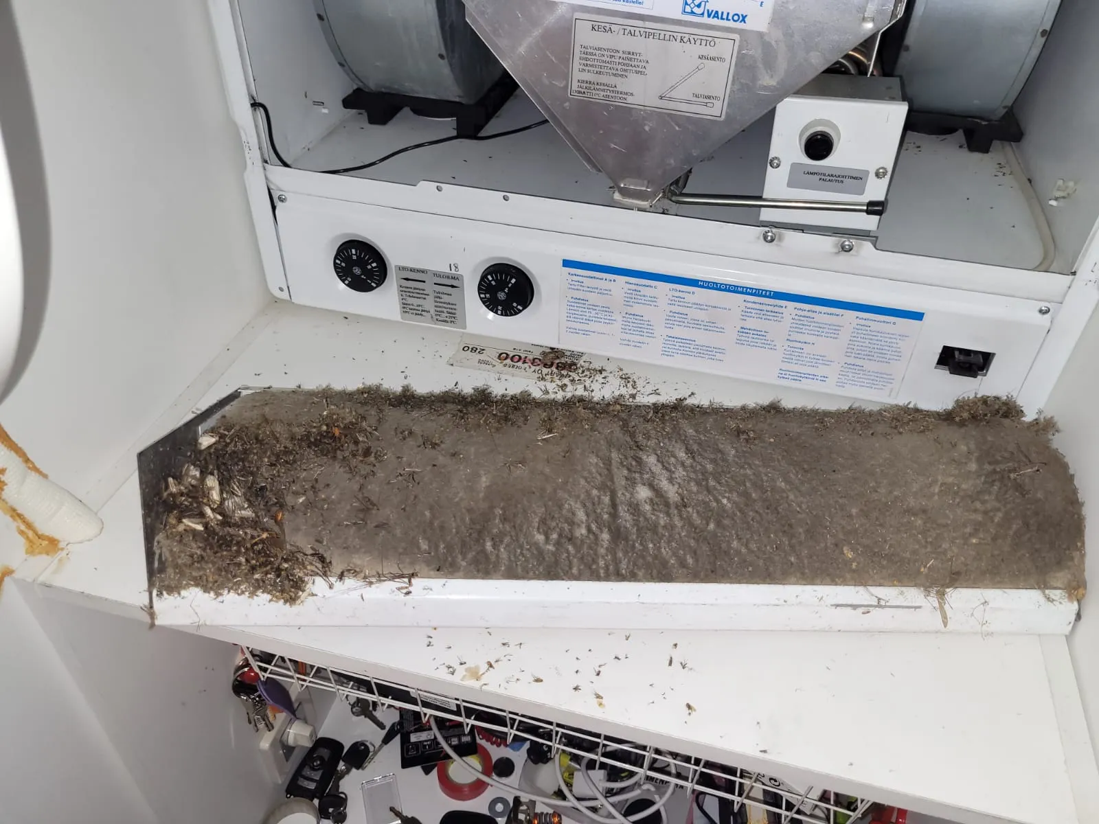 Likainen ilmansuodatin Vallox-ilmanvaihtokoneessa, täynnä pölyä ja roskia, valkoisessa laitteessa, jossa on säätönuppi ja huolto-ohjeita.