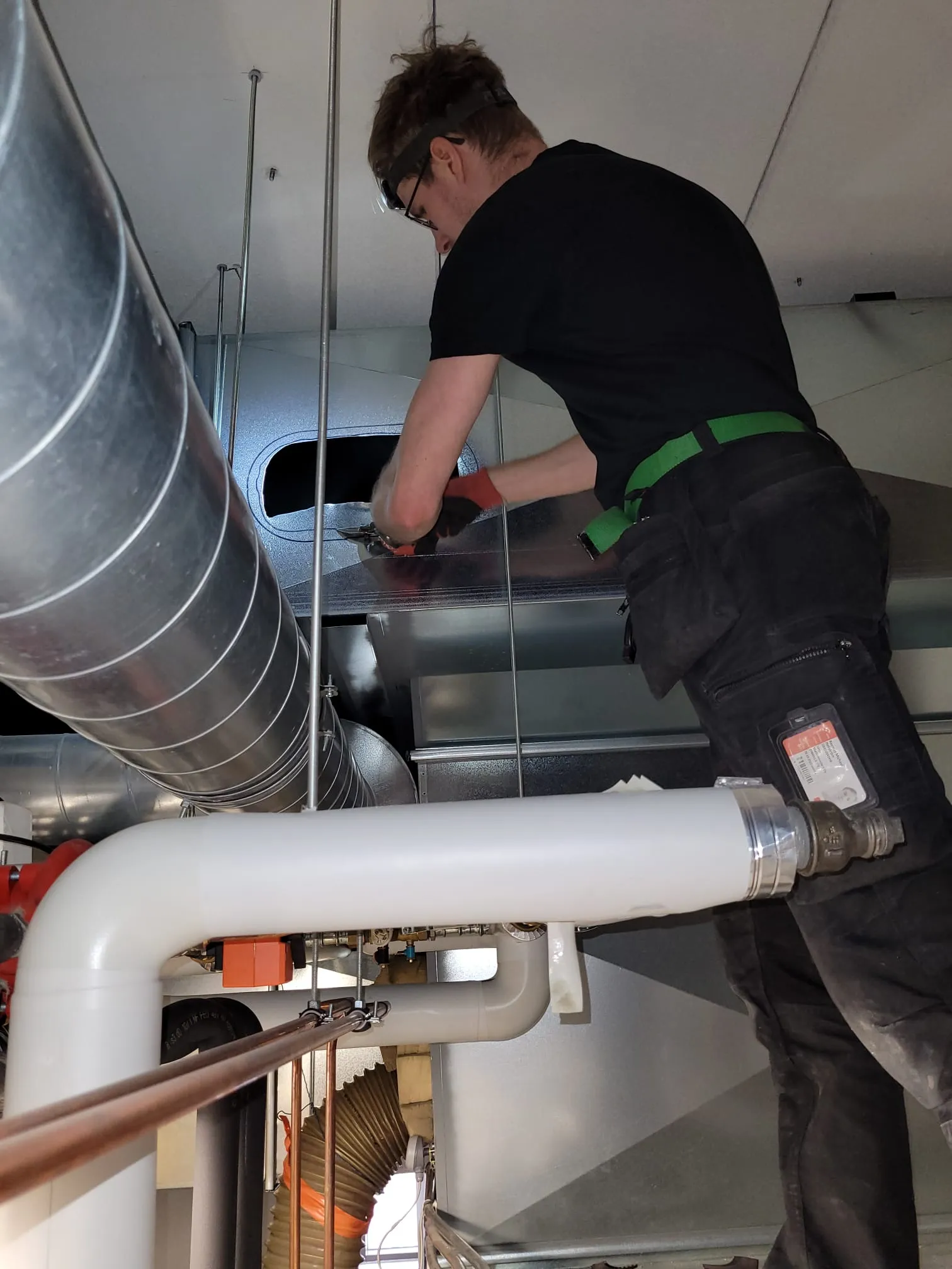 Kuvassa on mies, jolla on suojalasit ja joka työskentelee ilmastointikanavan parissa. Hänellä on yllään musta paita, tummat housut ja vihreä vyö. Kanavat ovat metallisia ja valaistus on kirkas.