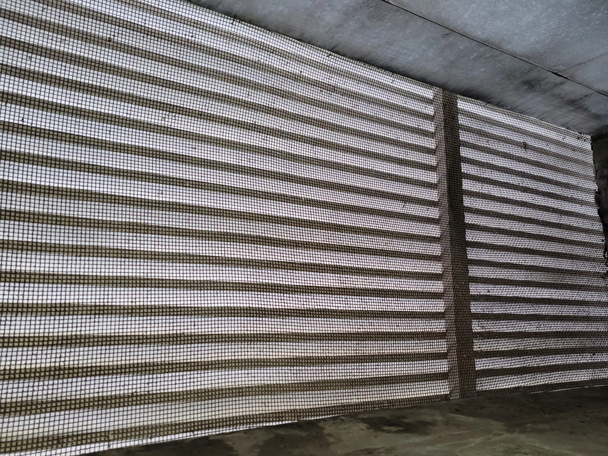 Kuva näyttää metalliverkon, jonka takana on vaakasuoria säleikköjä. Valo tulee läpi, luoden varjoja. Betonikatto näkyy yläreunassa.