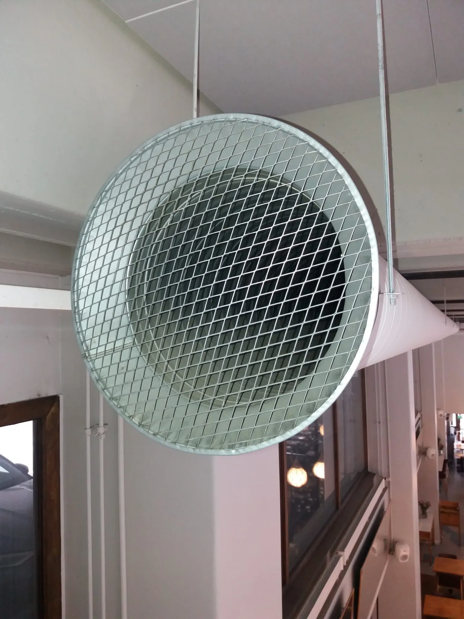 Metallinen ilmastointiputki roikkuu katosta, edessä suojaritilä. Vaaleat seinät ja kirkas valaistus.