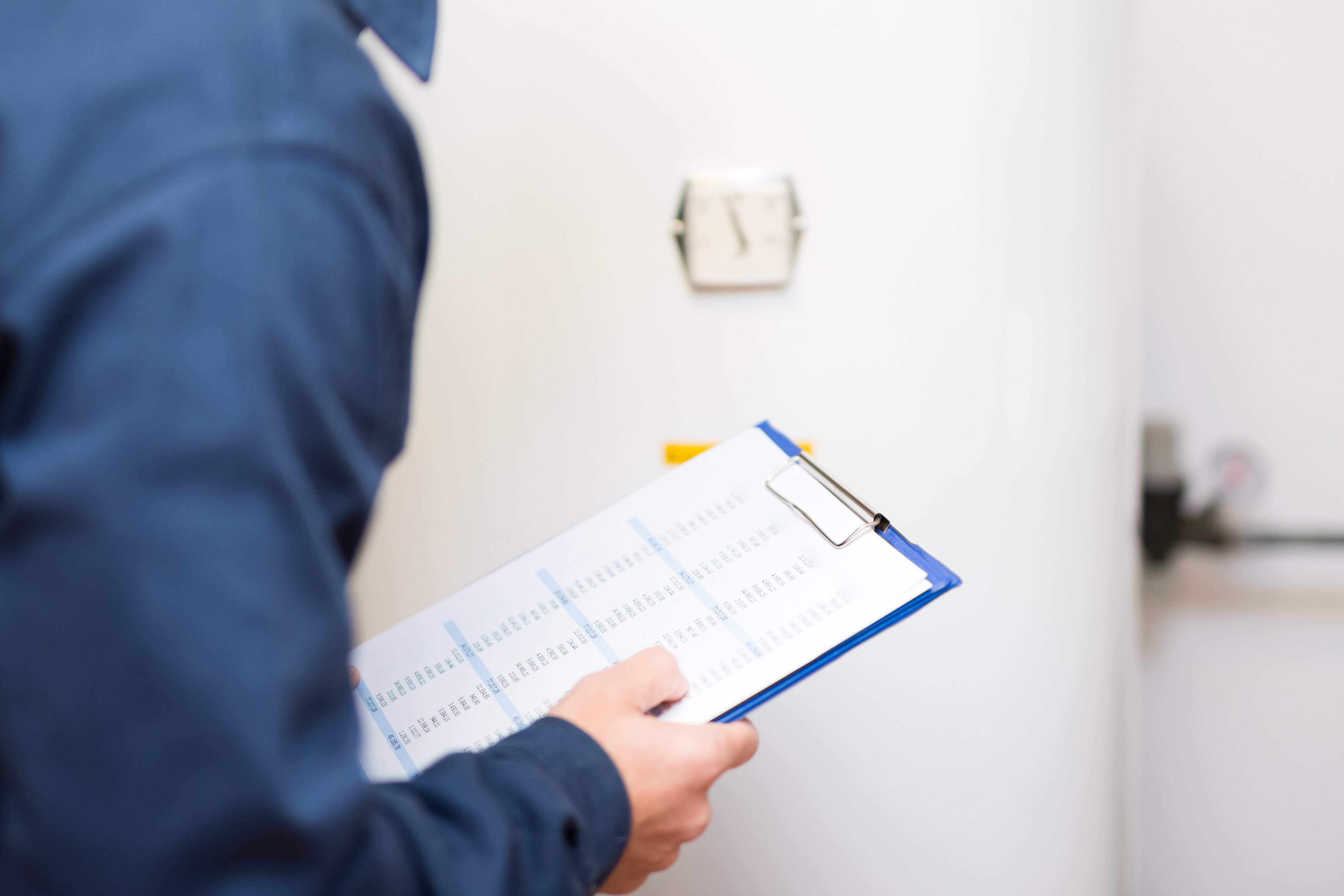 LVI-asentaja tarkistaa lämminvesivaraajaa, sininen paita, lehtiö kädessä, valkoinen tausta.