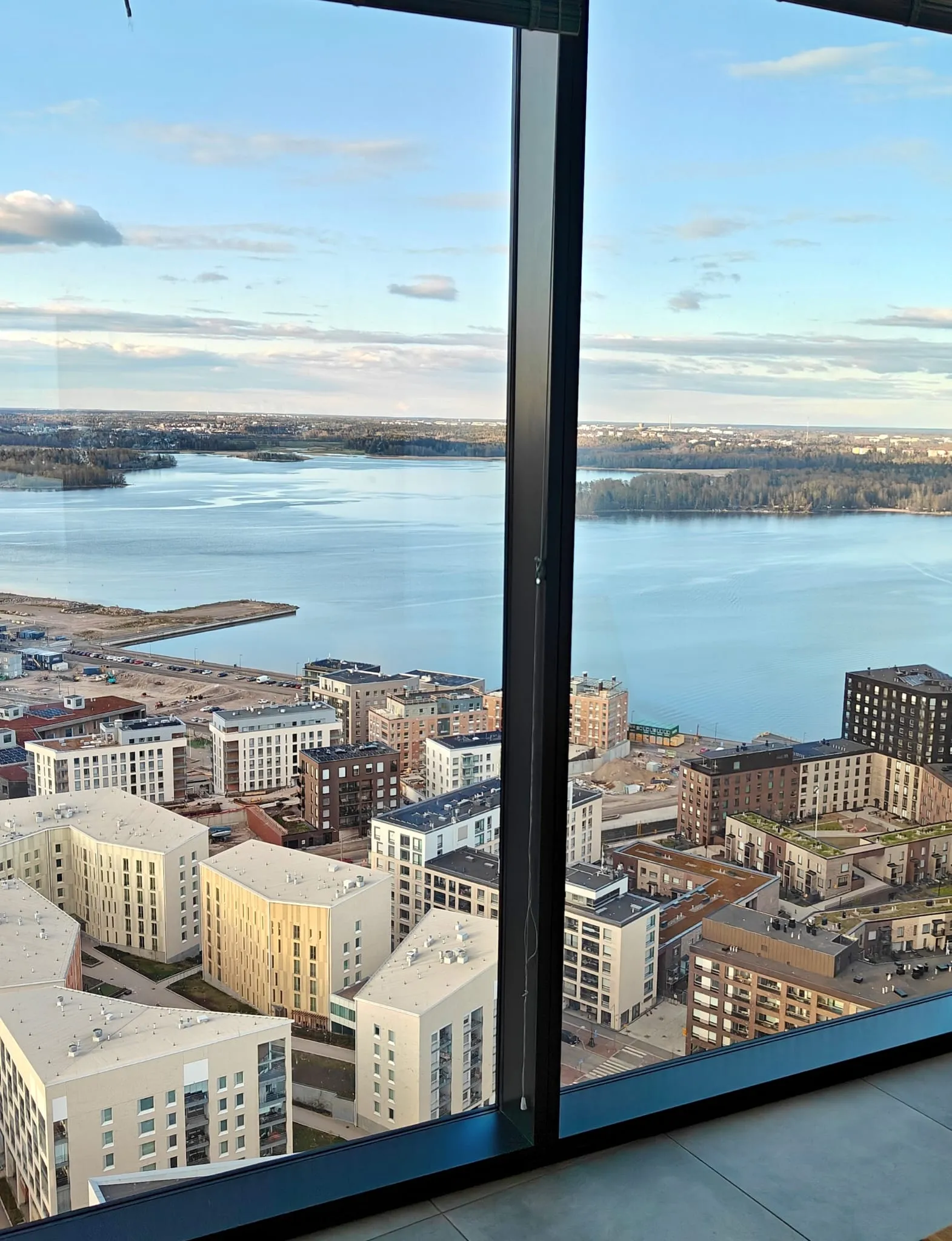 Näkymä korkealta rakennuksesta, jossa näkyy kaupunkimaisema, järvi ja taivas, kaikki ikkunan läpi. Rakennukset ovat pääosin vaaleita ja moderneja, ja järvi ulottuu horisonttiin asti. Taivaalla on pilviä ja valo on pehmeää.