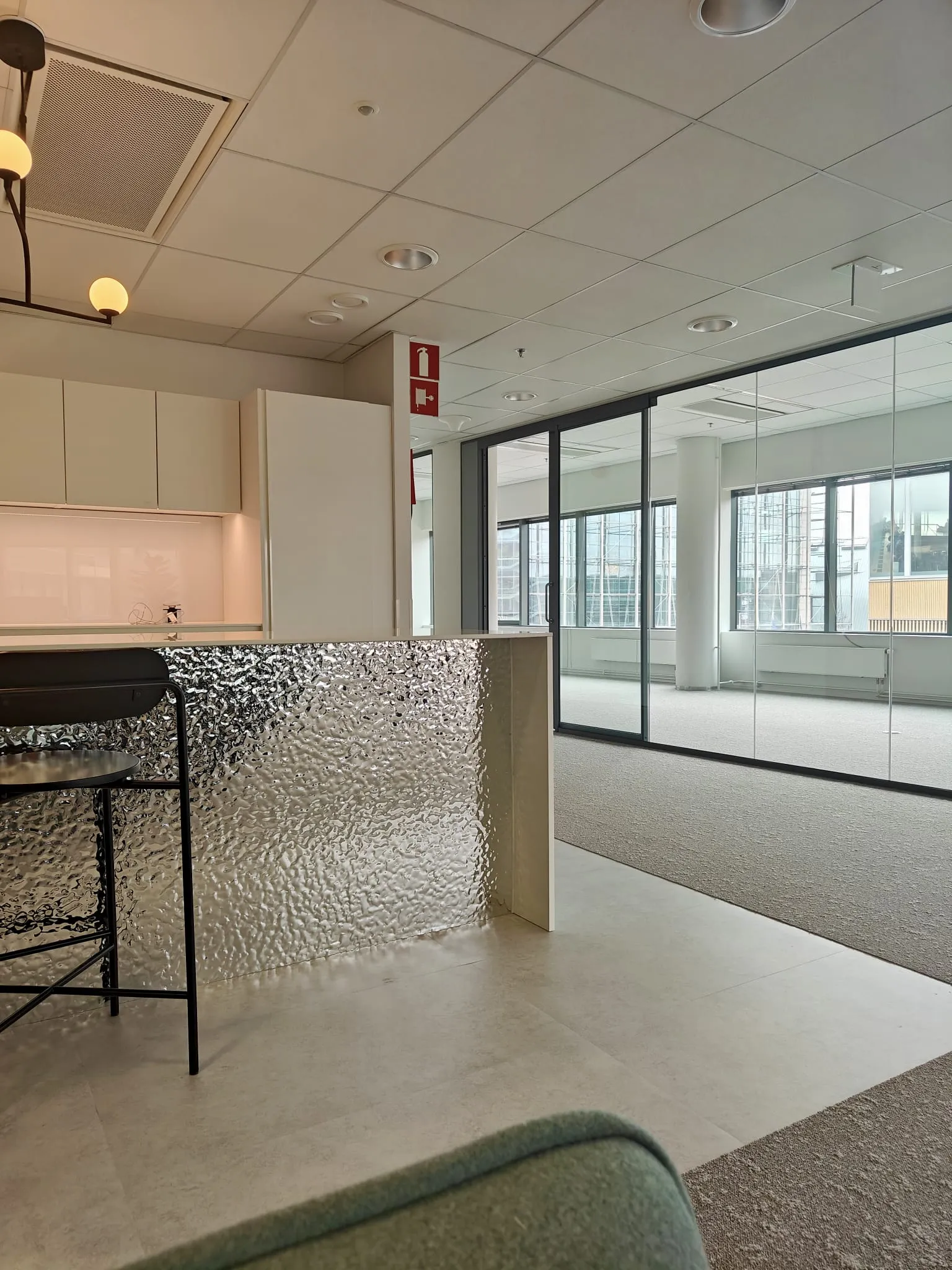 Sisätiloissa on keittiö, jossa on metallinen baaritiski ja musta tuoli, sekä lasiseinäinen toimisto, jossa on suuret ikkunat ja harmaa matto.