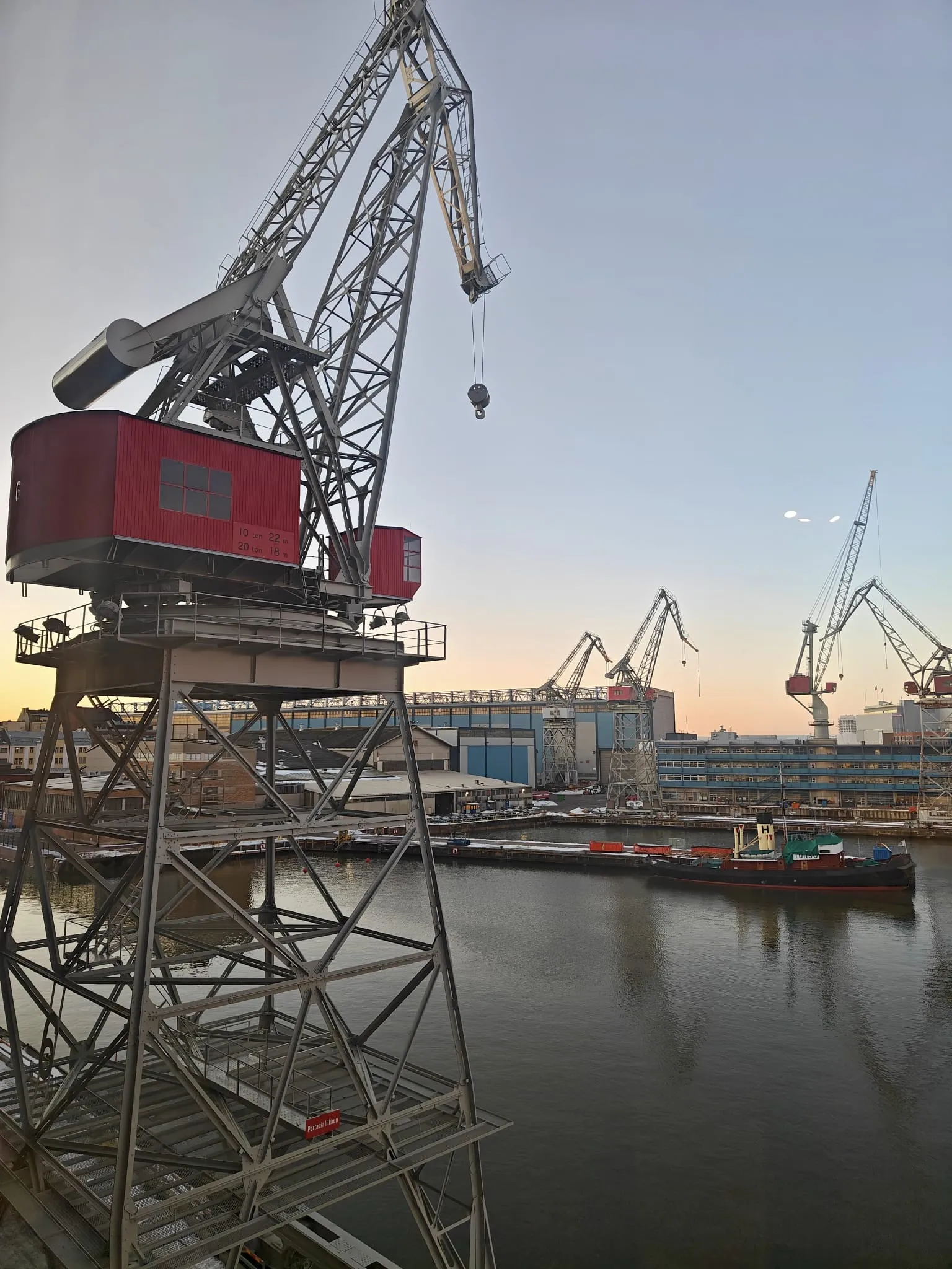 Satamanosturit kohoavat taivaalle, niiden punaiset ohjaamot erottuvat harmaasta teräksestä. Vesi kimmeltää auringonlaskun valossa, ja taustalla näkyy telakka rakennuksineen.