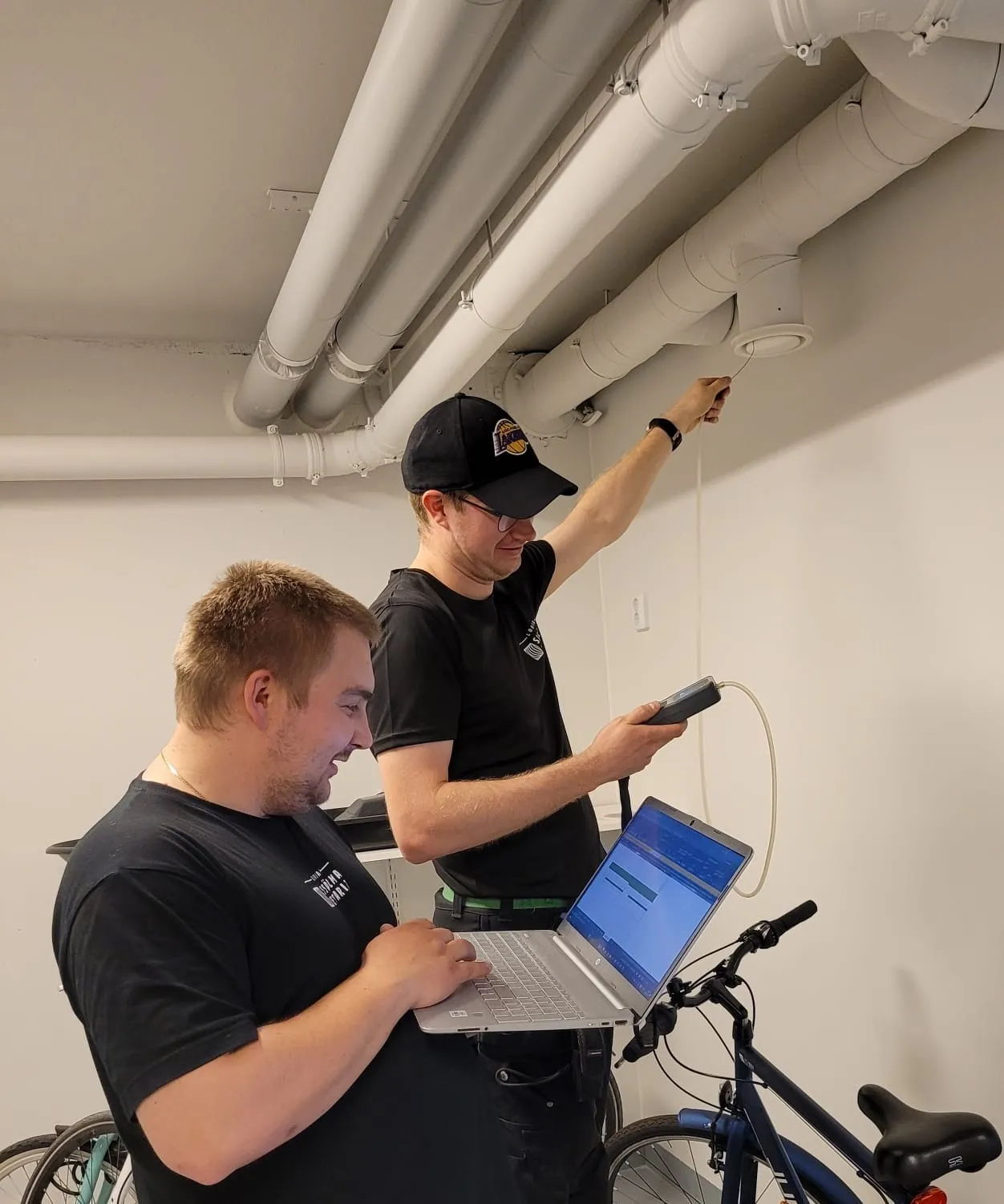 Kaksi miestä työskentelee sisätiloissa, toinen mittaa ilmanvaihtoputkea ja toinen käyttää kannettavaa tietokonetta, joka on kiinnitetty polkupyörään.