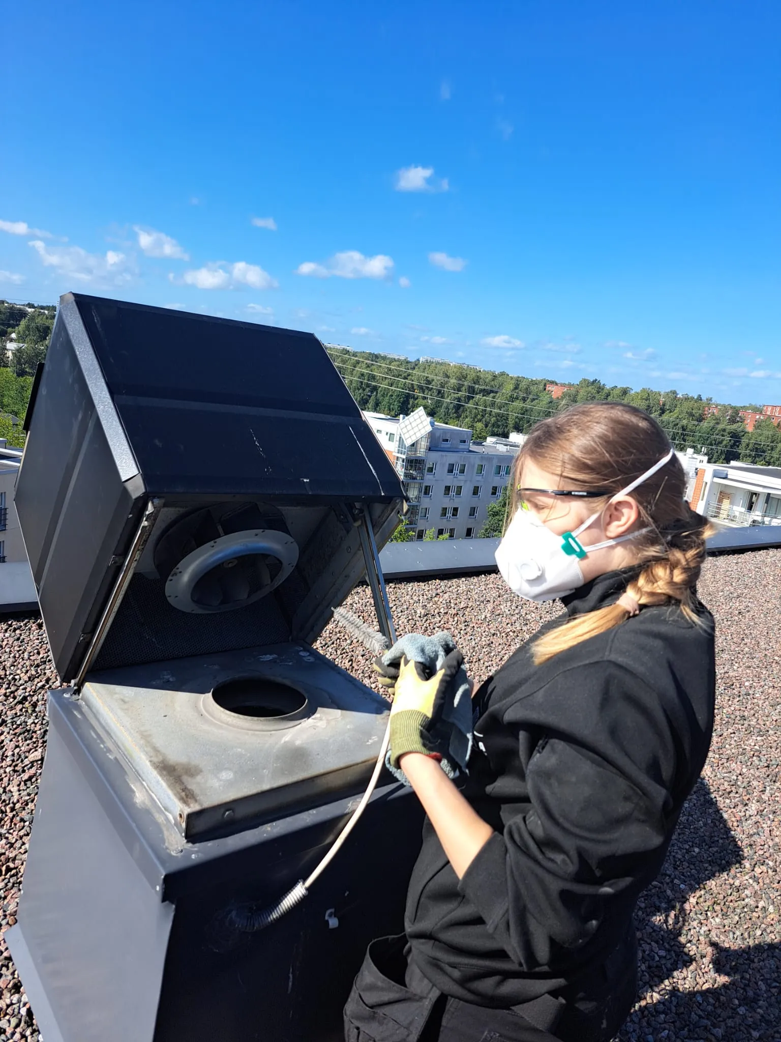 Tyttö suojavarusteissa puhdistaa ilmastointilaitetta katolla, aurinkoinen sää.